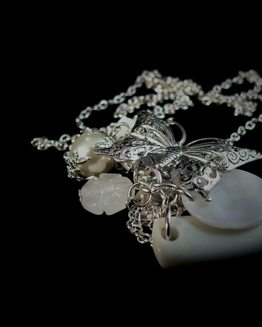 A silver filigree butterfly pendant among fine chains and carved shell blossoms, on black.